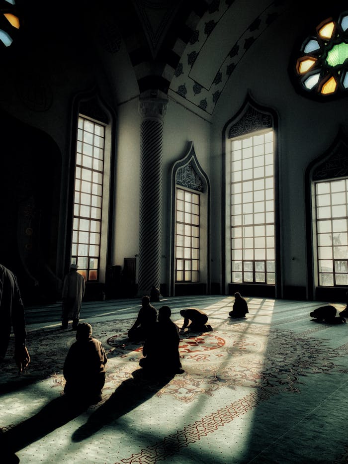 Silhouetted individuals praying inside a sunlit mosque with intricate architecture.